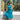 A woman wearing a vibrant green and blue patterned halter midi dress stands in front of a storefront window, holding out one side of the skirt to show its flow. The dress features a high-waisted design with a tie at the waist and a tiered ruffled hem.