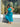 A woman wearing a vibrant green and blue patterned halter midi dress stands in front of a storefront window, holding out one side of the skirt to show its flow. The dress features a high-waisted design with a tie at the waist and a tiered ruffled hem.