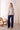 Woman wearing a patterned blouse and blue pants in a room with a white brick wall and wicker chair.