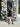 Woman in a black dress and boots standing between floral decorations on a building entrance.