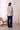Woman wearing a patterned blouse and dark pants standing in a room with a white brick wall and a wicker chair.
