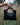 Black woven handbag held by a hand in front of tall grasses