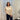 Woman wearing a beige sweater and dark pants standing in a room with white curtains and a lamp.