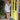 A woman stands on a storefront porch wearing a long-sleeve white and navy blue vertically striped mini dress with a V-neckline and matching striped socks. The background features a bright yellow-green door, potted plants, and a concrete floor.