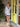 A woman stands on a storefront porch wearing a long-sleeve white and navy blue vertically striped mini dress with a V-neckline and matching striped socks. The background features a bright yellow-green door, potted plants, and a concrete floor.