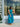 A woman wearing a sleeveless teal and blue floral midi dress stands in front of a clothing store window display.