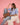 Woman sitting on a chair holding a brown woven handbag against a pink background